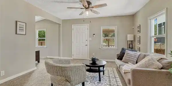 Image 4: Bright and spacious living area featuring neutral wall finishes, ceiling fan with brushed metal accents, carpet flooring throughout, and multiple windows allowing natural light