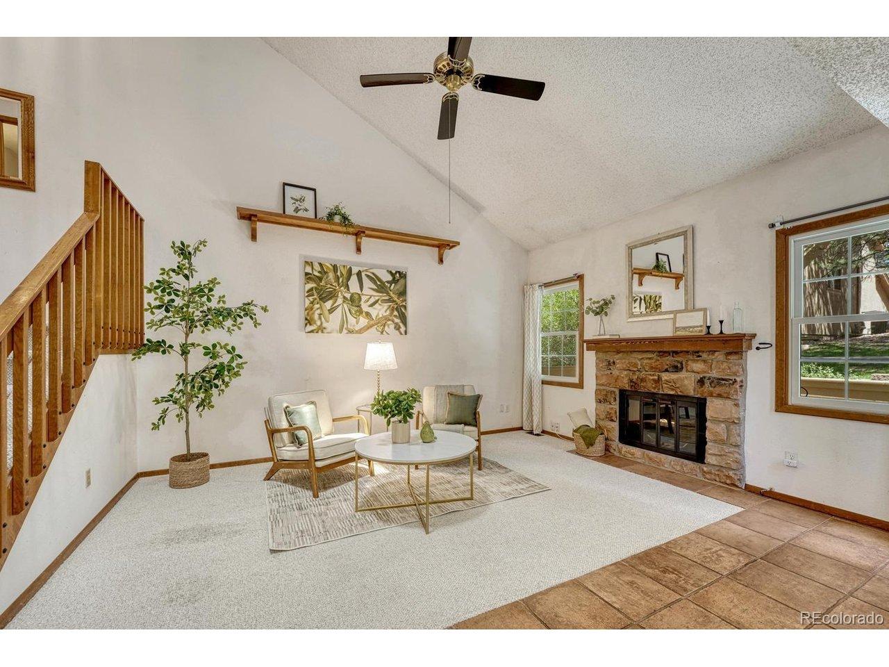 Image 4: Bright, inviting vaulted living room featuring neutral carpeting, a ceiling fan, wall shelf, & cozy wood-burning fireplace with a classic brick surround to cozy up on chilly evenings.