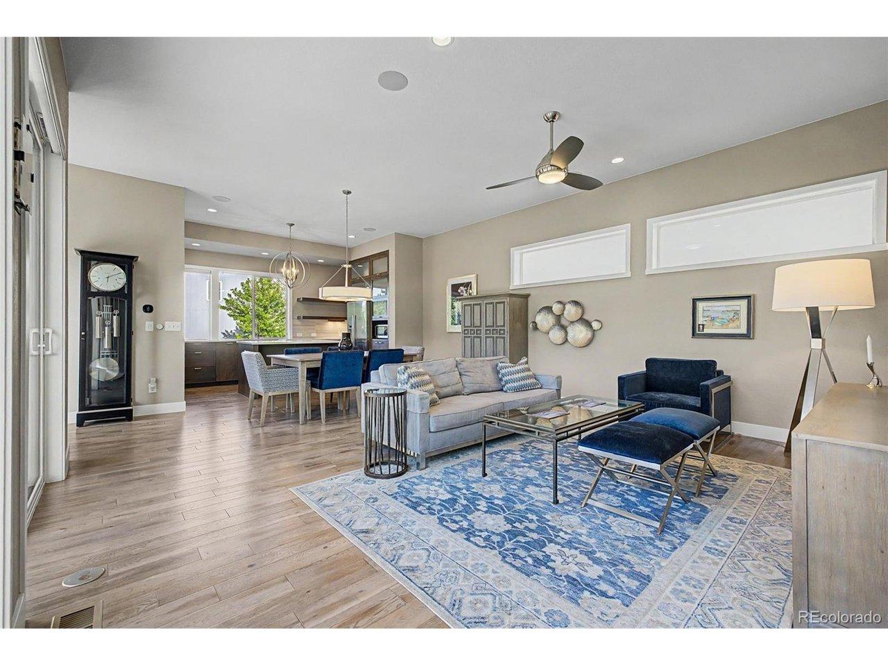 Image 4: Spacious open-concept great room with soaring ceilings, wide-plank hardwood floors, and a cohesive designer color palette. Flows effortlessly into the dining and kitchen areas, anchored by a bold blue area rug and statement ceiling fan.