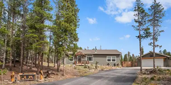 Image 3: Wind your way up a paved drive flanked by towering pines, and the home reveals itself just where the trees open up. Blue skies overhead, wood beam accents on the garage, a covered front porch waiting at the end - this is the kind of arrival that makes ever