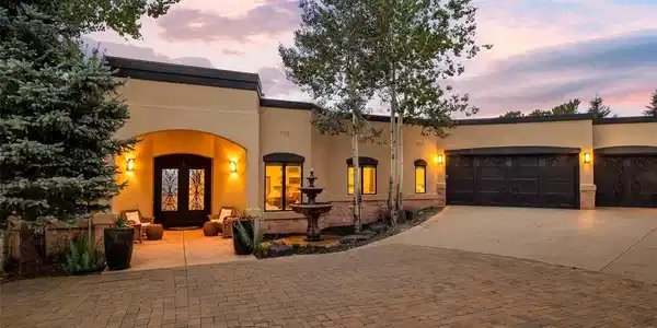 Image 4: The welcoming front elevation glows at dusk with warm lighting, a tiered stone fountain, mature aspens, and elegant wrought-iron entry and garage doors.
