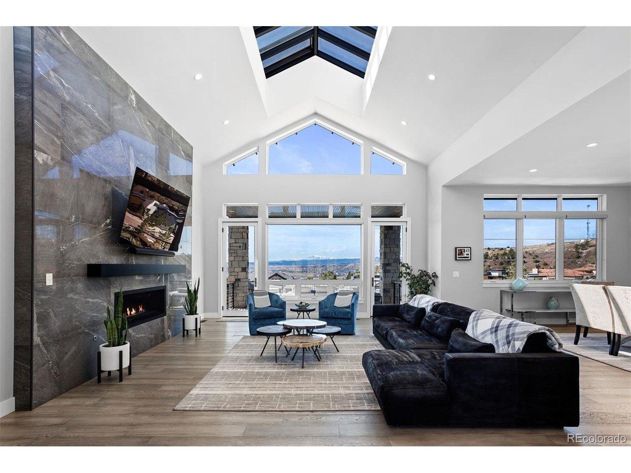 Image 3: Twenty-foot vaulted great room with custom ridge skylight, floor-to-ceiling windows with unobstructed Pikes Peak views, linear fireplace with Italian marble surround, and two doors opening to the covered deck.