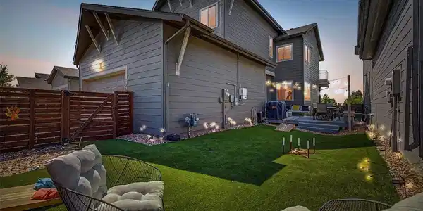 Image 3: Magical twilight view of the fully fenced backyard showcasing lush artificial turf, landscape lighting, lawn game area, and a beautifully lit composite deck with string lights and outdoor dining. The detached garage, upper balcony, and glowing interior win Image 3: Magical twilight view of the fully fenced backyard showcasing lush artificial turf, landscape lighting, lawn game area, and a beautifully lit composite deck with string lights and outdoor dining. The detached garage, upper balcony, and glowing interior win