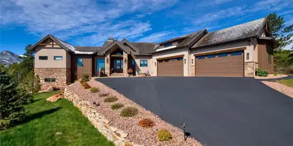Image 2: Built for Colorado living. Expansive side elevation featuring a 4-car attached garage with custom carriage-style doors, mixed stone and stucco facade, multilevel landscaping, and direct mountain views from the driveway.