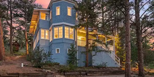 Image 2: Rising among the pines, this three-story blue beauty glows warm from within as evening falls. A wraparound porch invites you to sit back and soak in the mountain air, while a charming staircase winds up through the trees to greet you at the door. This isn' Image 2: Rising among the pines, this three-story blue beauty glows warm from within as evening falls. A wraparound porch invites you to sit back and soak in the mountain air, while a charming staircase winds up through the trees to greet you at the door. This isn'