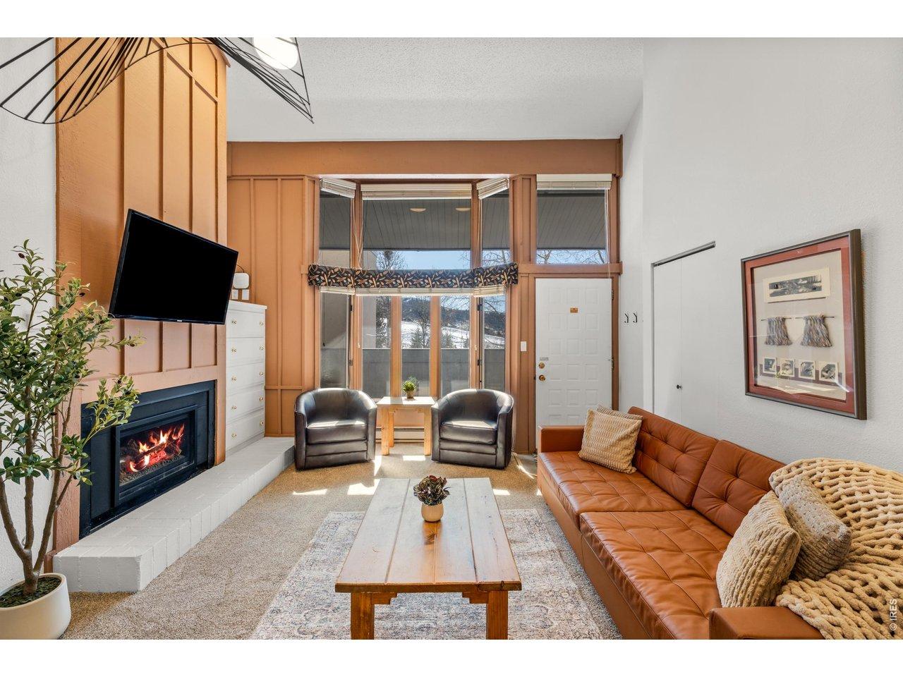 Image 3: Relax by the fireplace in this bright, inviting living room with large windows and cozy seating. Perfect for morning coffee, apr s-ski drinks, or winding down with a movie after a day in the mountains.