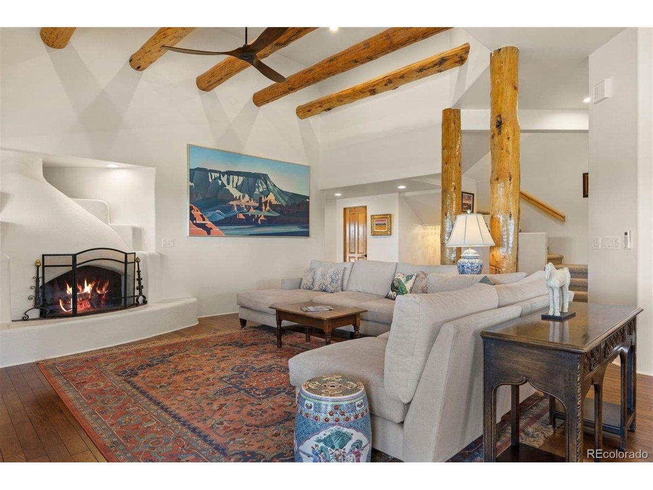 Image 4: The great room showcases a 14-foot ceiling with hand-hewn log beams, natural column supports, and a kiva-style wood-burning fireplace framed by a traditional banco bench. Image 4: The great room showcases a 14-foot ceiling with hand-hewn log beams, natural column supports, and a kiva-style wood-burning fireplace framed by a traditional banco bench.