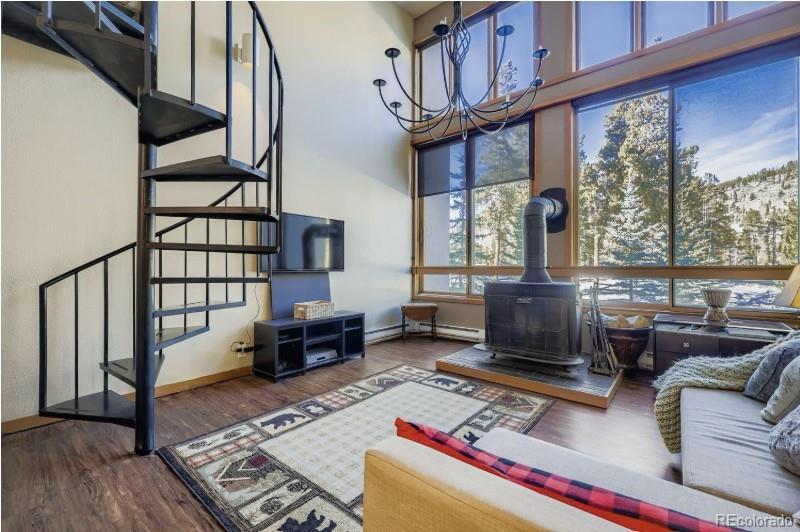 Image 1: Living room view from kitchen/dining nook. Floor to ceiling windows. Large windows are sliders with screens! Wood burning stove. Wood provided by HOA. Spiral staircase to primary bedroom & full bathroom.